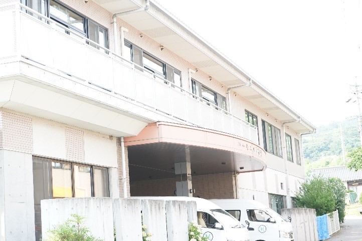 介護施設の建物外観。白と煉瓦造りの複数階建て。駐車場に車が停まっている。